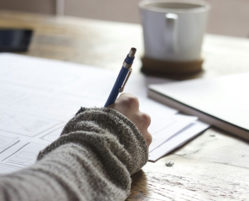 person writing on brown wooden table near white ceramic mug